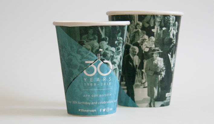 A small and large take-away cup featuring the 30 years brand identity with a historic photograph of the Queen and Prime Minister Bob Hawke from the day the building opened to the public.